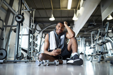 Happy young sportsman with smartphone sitting on the floor in fitness center and enjoying music at break