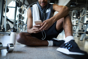 African-american sportsman with smartphone sitting on the floor, relaxing after workout and listening to music in earphones