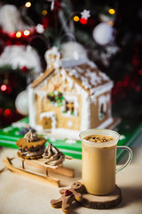 Close-up mug with coffe and milk on a wooden table, gingerbread house and christmas lights and decorations on bokeh