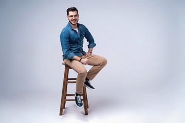 Full length portrait of an attractive young man in jeans shirt sitting on the chair over grey...
