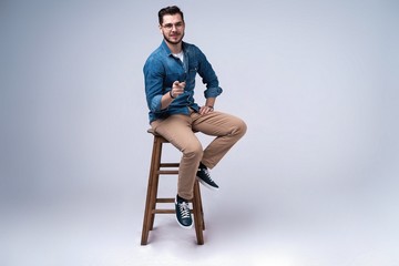 Full length portrait of an attractive young man in jeans shirt sitting on the chair over grey...