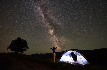 Active happy woman backpacker having a rest at night camping in mountains. Female standing beside illuminated tent with hands raised up, enjoying view of amazing night sky full of stars and Milky way