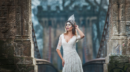 Obraz premium Lovely young lady wearing elegant white dress enjoying the beams of celestial light and snowflakes falling on her face. Pretty brunette girl in long wedding dress posing on a bridge in winter scenery