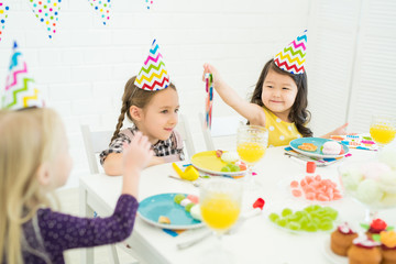 Cheerful Asian child with curly hair wearing colorful party hat sitting at table with food and giving paper napkin to birthday girl while congratulating her at birthday party