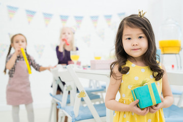 Content attractive Asian little queen with gift box wearing yellow dress standing at dining table and looking at camera while her friends blowing party horns in background.
