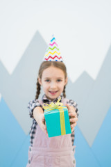 Positive cute girl in party hat standing against wall with mountains drawing and giving blue gift box to camera