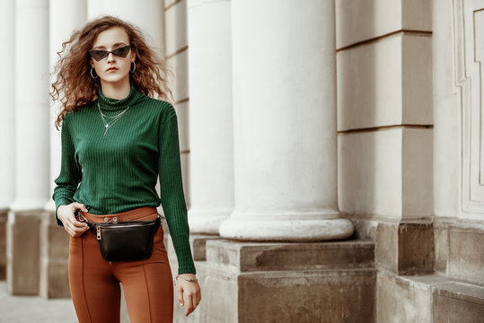 Outdoor fashion portrait of young lady wearing sunglasses, green turtleneck, brown trousers, leather belt bag, necklace, bracelet, posing in street of european city. Copy, empty space for text
