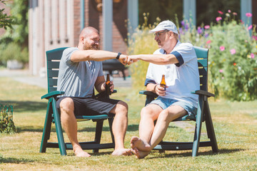 Two men drinking beer together and making power five gesture in summer sunny garden (Netherlands -...