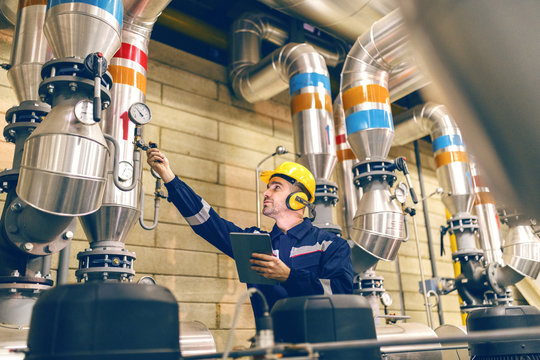 Young Caucasian worker in protective suit tightening the valve and using tablet while standing in heating plant.