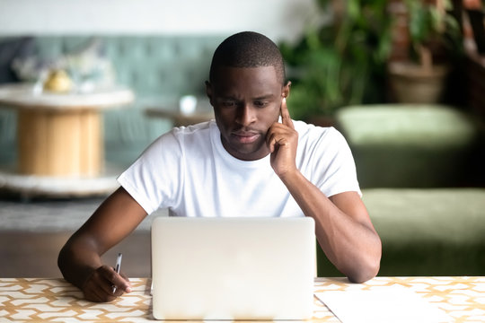 Focused African American man using laptop in cafe, serious black student looking at computer screen, thinking about online project, task, reading e-book, news, journalist writing, think about article