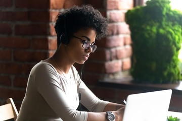 Focused African American woman wearing headphones using laptop, making writing notes, serious...