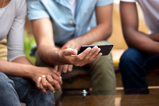 Close up man holding phone in hands, show photos, using mobile device with diverse friends, multiethnic people looking at smartphone screen, watching video in social network, sitting together