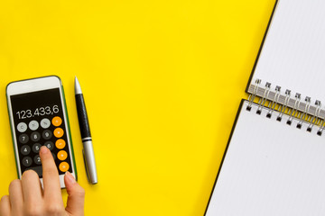 Accounting, financial concept, flat lay or top view of pen, smart phone with calculator with white notepad on vivid yellow background table with blank copy space, math, cost, tax or investment calcula