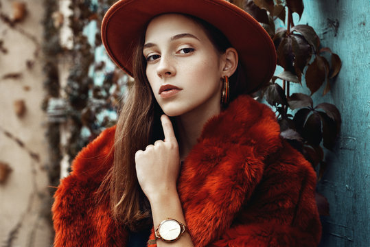 Outdoor close up fashion portrait of young beautiful confident woman wearing trendy orange faux fur coat, hat, hoop earrings, wrist watch, posing in street of european city. Copy, empty space for text