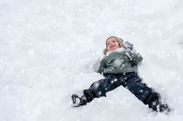 Fototapeta na wymiar Little boy lying on his back in the snow and having fun while snow is falling