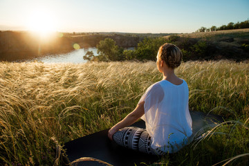 Naklejka na ściany i meble Girl meditates sitting on a beautiful meadow at sunset