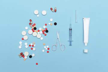 Scattered pills, lying scissors, syringe and tube of ointment on a blue background