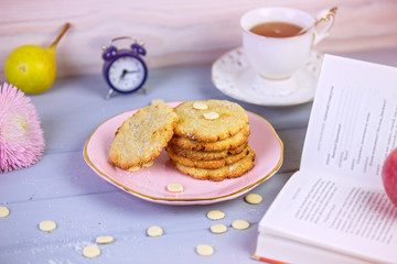 Freshly baked coconut cookies with white chocolate on a beautiful plate, on a gray wooden background, surrounded by a cup of tea, clock, fruit, books. Still life in a cafe