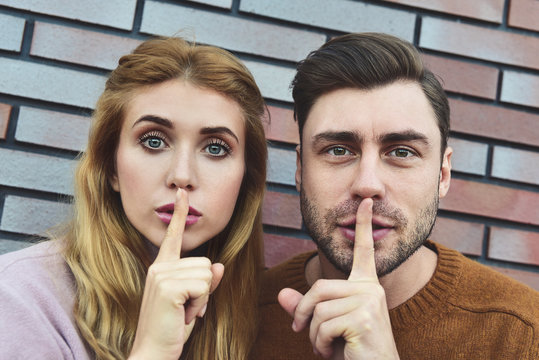 Shh, Dont Tell To Anybody This Private Information. Surprised Caucasian Couple Make Shush Gesture With Surprised Expressions, Ask Not Spread Gossips About Colleagues, Isolated Over Brick Background.