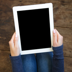The girl sits on a wooden floor and holds a tablet with a blank screen in her hands, top view. Closeup.