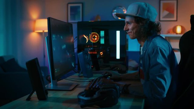 Professional Gamer Playing First-Person Shooter Online Video Game on His Powerful Personal Computer. Room and PC have Colorful Neon Led Lights. Young Man is Wearing a Cap. Cozy Evening at Home.