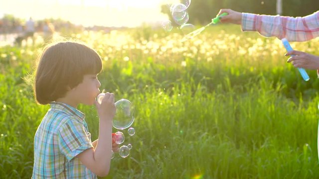 Close up portrait of happy little cute boy blowing, having fun with soap bubbles in park. Little child playing outside on summer sunny day in slowmo. Happy holiday, vacation, childhood, summertime
