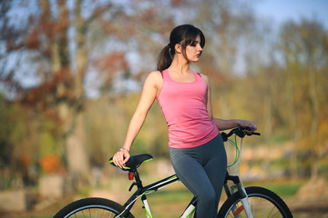 Naklejka premium Young beautiful girl in sportswear stands next to a bicycle on the street in the park in autumn.