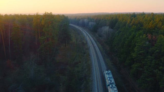 Aerial following view of the freight train carrying tank cars with oil moving through the autumn forest at sunset