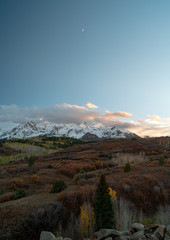 Fototapeta premium The sun begins going down over partly cloud hidden snow capped mountain ranges above autumn colored farm land and forests