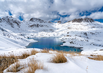 A very deep blue lake sits in a valley between snow capped mountains on a partly cloudy day