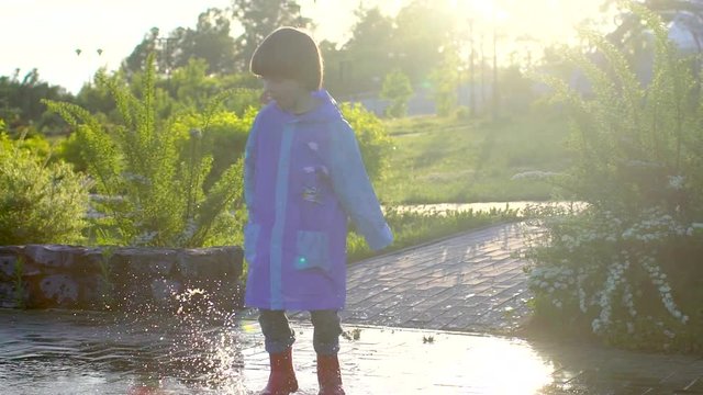 Happy cute little boy chid running playing having fun jumping through puddles in raincoat and rubber boots. laughing and smiling boy in park. rain slush spring summer autumn bad weather puddle cold