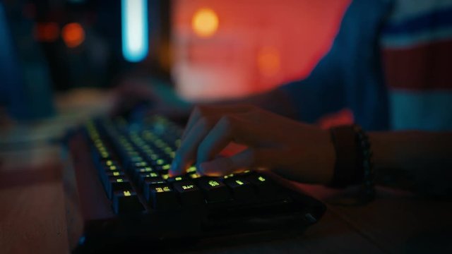 Close Up Hands Shot Showing a Gamer Pushing the Keyboard Buttons while Playing an Online Video Game. Keyboard Led Lights Change Color in Rainbow Spectrum. Gamer is Wearing a Bracelet. Room is Dark.