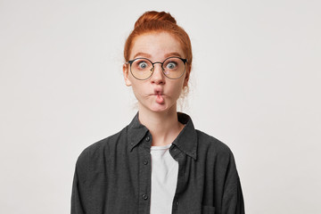 Red-haired girl portrays a fish with her lips looking through glasses with wide eyes. Woman doing fish face isolated on white background - obrazy, fototapety, plakaty