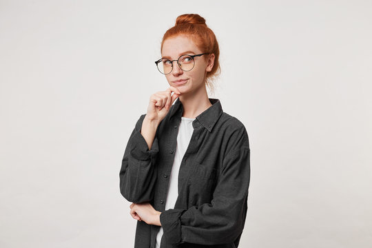 Portrait of young red -hair female in black men's shirt, keeps arm on chin and looks up, ponders the idea, consider different variants, thinking what do to solve the problem, over white background