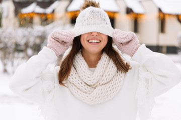 Outdoor close up portrait of young beautiful happy smiling girl wearing white knitted, scarf and gloves. The model jokes and closes the eyes with hat in park with Christmas lights. 