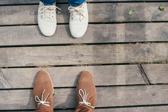 Guy and girl standing in front of each other wearing white sport shoes and brown casual shoes. Couple walking outdoors in the afternoon. No face.