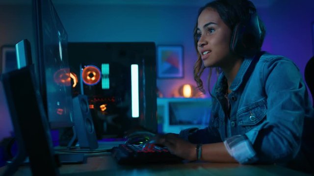 Pretty and Excited Black Gamer Girl in Headphones is Playing and Winning in First-Person Shooter Online Video Game on Her Computer. Room and PC have Colorful Neon Led Lights. Cozy Evening at Home.