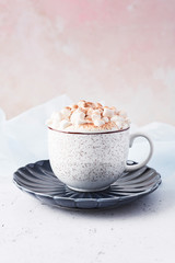 Hot chocolate or cocoa with whipped cream and marshmallow candy sprinkled with cinnamon or cocoa powder on white marble table over blue silk and pink background with copy space for text.