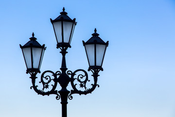 Fototapeta na wymiar Silhouette of a black street lamp against a blue sky, lamppost with three wrought iron lamps with glass inserts.