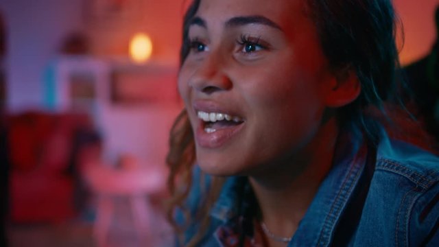 Close Up Portrait of a Beautiful Excited Black Girl Watching an Action Video on a Computer. She Has Dark Hair and Brown Eyes. Screen Adds Reflections to Her Face. Cozy Room is Lit with Warm Red Light.
