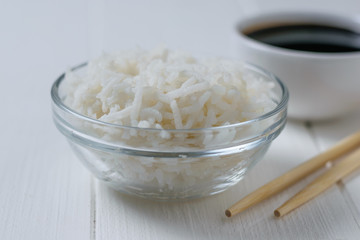 Glass bowl with long grain rice and wooden spoon on white wooden table.