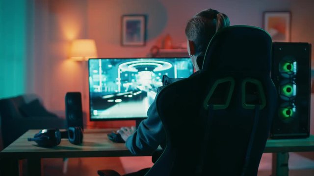 Back Shot of a Gamer Playing First-Person Shooter Online Video Game on His Powerful Personal Computer. Room and PC have Colorful Neon Led Lights. Cozy Evening at Home.