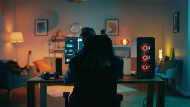 Back Shot of a Gamer Playing and Winning in First-Person Shooter Online Video Game on His Powerful Personal Computer. Room and PC have Colorful Neon Led Lights. Cozy Evening at Home.