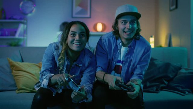 Excited Black Gamer Girl and Young Man Sitting on a Couch and Playing Video Games on Console. They Plays with Wireless Controllers. Cozy Room is Lit with Warm and Neon Light.