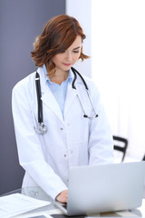 Happy doctor woman at work. Portrait of female physician using laptop computer while standing near reception desk at clinic or emergency hospital. Medicine concept