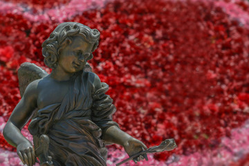 Close cherub Cupid portrait. Little boy angel sculpture holds the rose in left hand with blurry bokeh background from beautiful red and pink roses. Image for Valentine's day or sweet love concept