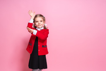 Diligent little girl in bright red jacket and black school dress smiling and holding up hand while standing on pink background