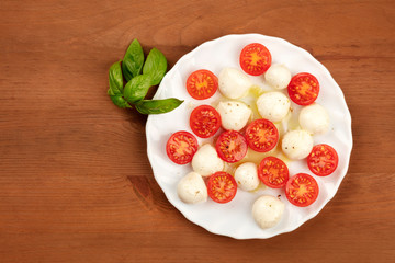 A photo of a plate of Italian Caprese salad with Mozzarella cheese, cherry tomatoes, and basil leaves, shot from above on a dark rustic wooden background with a place for text