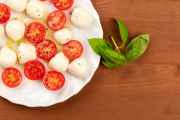 A closeup photo of a plate of Italian Caprese salad with Mozzarella cheese, cherry tomatoes, and basil leaves, shot from above on a dark rustic wooden background with a place for text