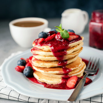 Stack of pancakes with berry sauce, blueberry, strawberry and cranberry. Cup of coffee and cream on background. Square crop
